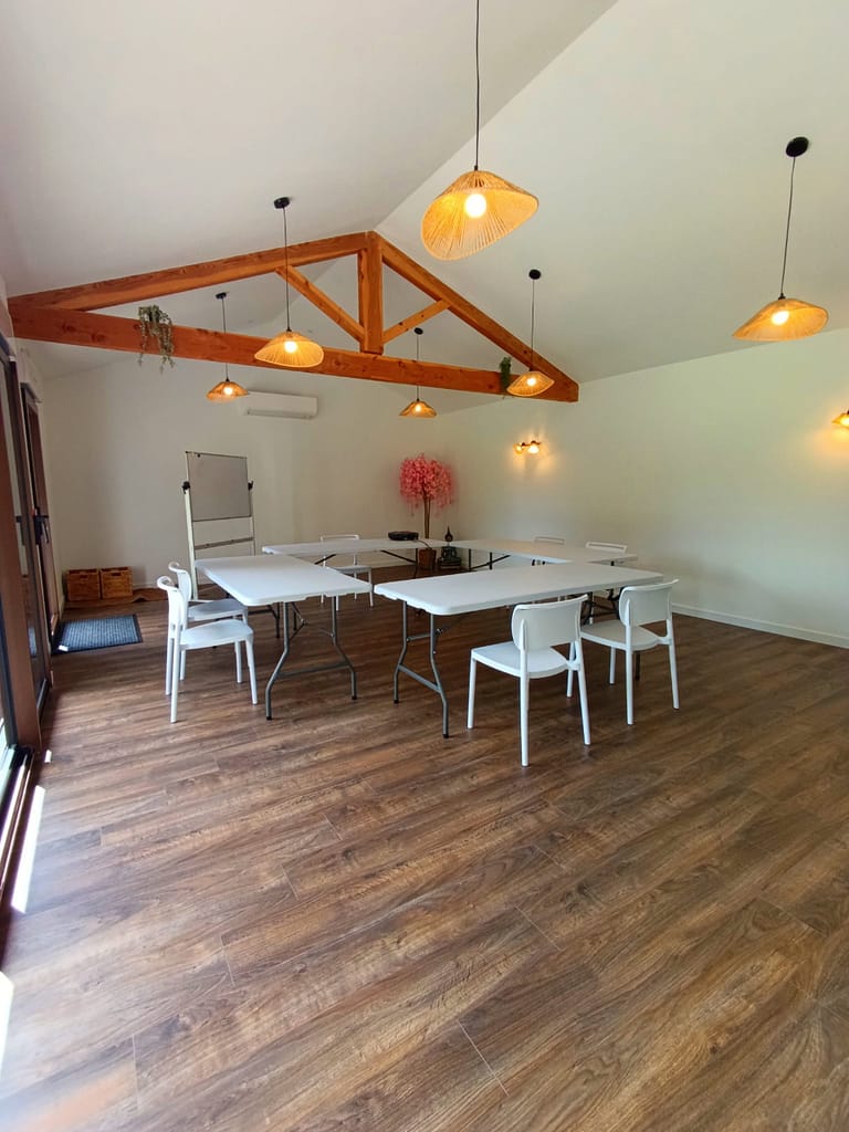 Cette photo montre une salle de réception lumineuse et conviviale, idéale pour les évènements d'entreprise, les séminaires, les ateliers bien-être ou le coworking. L'espace comprend un vidéoprojecteur, de grandes tables modulables, des chaises confortables, ainsi qu'un coin détente avec canapés. Une cuisine entièrement équipée permet d'organiser des pauses cafés ou des repas sur place. L'ambiance chaleureuse et professionnelle crée un cadre propice à la collaboration et à la créativité