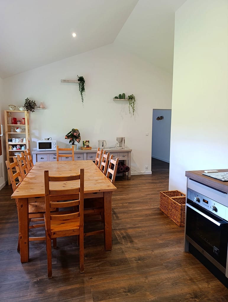 Cette photo montre une salle de réception lumineuse et conviviale, idéale pour les évènements d'entreprise, les séminaires, les ateliers bien-être ou le coworking. L'espace comprend un vidéoprojecteur, de grandes tables modulables, des chaises confortables, ainsi qu'un coin détente avec canapés. Une cuisine entièrement équipée permet d'organiser des pauses cafés ou des repas sur place. L'ambiance chaleureuse et professionnelle crée un cadre propice à la collaboration et à la créativité