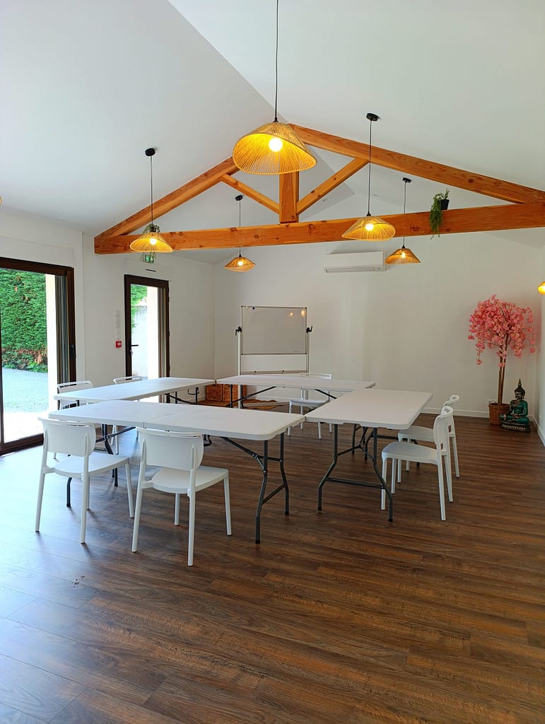 Cette photo montre une salle de réception lumineuse et conviviale, idéale pour les évènements d'entreprise, les séminaires, les ateliers bien-être ou le coworking. L'espace comprend un vidéoprojecteur, de grandes tables modulables, des chaises confortables, ainsi qu'un coin détente avec canapés. Une cuisine entièrement équipée permet d'organiser des pauses cafés ou des repas sur place. L'ambiance chaleureuse et professionnelle crée un cadre propice à la collaboration et à la créativité